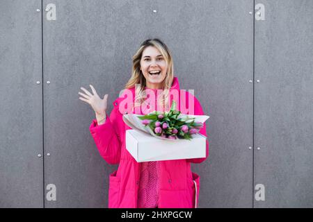 Portrait of smiling surprised blond woman, getting pink fresh bouquet of tulip flowers and present box. Delivery gift Stock Photo