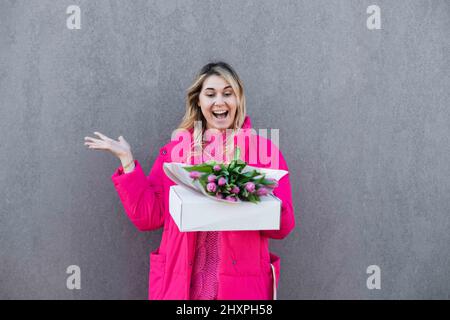 Portrait of laughing glad surprised blond woman, getting pink fresh bouquet of tulip flowers and present box. Gray view Stock Photo