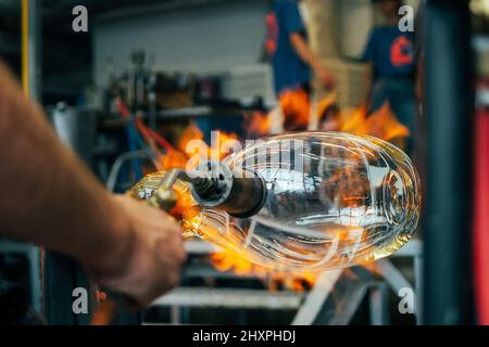 Close up of a glassblower artisan shaping the hot molten glass at strong fire inside a workshop. Manual glass processing by the craftsmen inside a gla Stock Photo