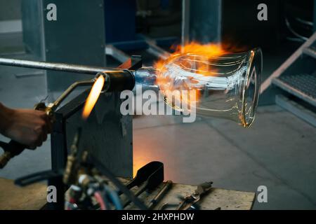 Close up of a glassblower artisan shaping the hot molten glass at strong fire inside a workshop. Manual glass processing by the craftsmen inside a gla Stock Photo