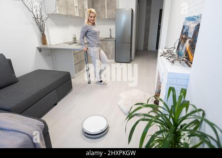 young woman using automatic vacuum cleaner to clean the floor, controlling smart machine housework robot Stock Photo