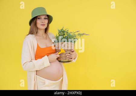 Portrait of modern confident pregnant smiling woman in green hat, orange top with plant pot. Cultivation and care flowers Stock Photo