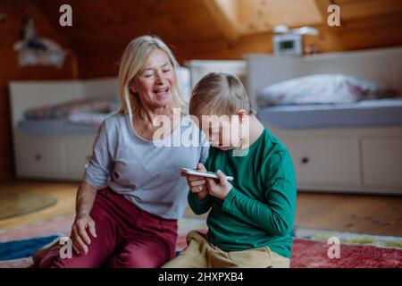 Child with Down syndrome sitting on floor and using tablet with grandmother at home. Stock Photo