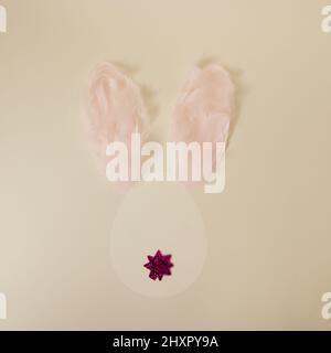 Pretty egg shaped Easter bunny from behind with ears made from pastel pink feathers and a decorative bow tail on yellow minimal background Stock Photo
