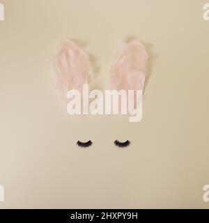Girl Easter bunny with ears made from pastel pink feathers and dark black eye lashes on yellow minimal background Stock Photo