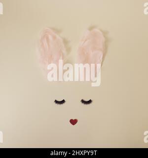 Girly Easter bunny with ears made from pastel pink feathers, heart shaped nose and dark black eye lashes on yellow minimal background Stock Photo