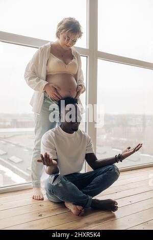Young happy african man meditates next his caucasian pregnant woman against window. Interracial marriage concept. Stock Photo
