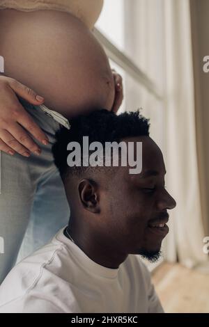 Closeup view of face of happy african man next belly his caucasian pregnant woman. Interracial marriage concept. Stock Photo