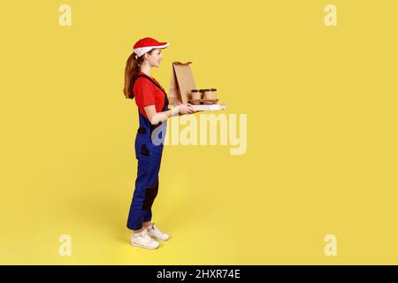 Side view portrait of courier woman holding coffee and boxes with pizza, offering drinks and food delivery, wearing overalls and red cap. Indoor studio shot isolated on yellow background. Stock Photo