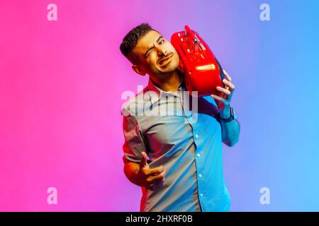 Funny handsome man in shirt listening to music holding red tape recorder on shoulder, winking at camera and showing thumb up. Indoor studio shot isolated on colorful neon light background. Stock Photo