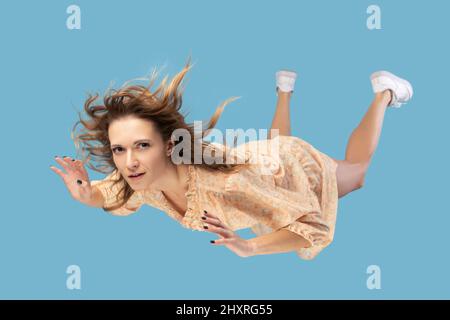 Beautiful young woman levitating in mid-air, falling down and her hair messed up soaring from wind, model flying hovering with dreamy peaceful expression. indoor shot isolated on blue background Stock Photo