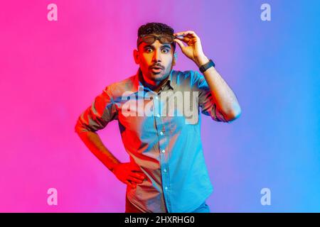 Portrait of astonished surprised man in shirt standing raised glasses and looking at camera with big eyes and shocked expression. Indoor studio shot isolated on colorful neon light background.