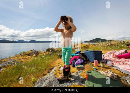 A young girl looks through binoculars on a mossy rock overlooking bay Stock Photo