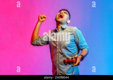 Portrait of man in shirt holding hand red joystick pad showing yes gesture sincerely rejoicing, victory in online virtual game. Indoor studio shot isolated on colorful neon light background. Stock Photo