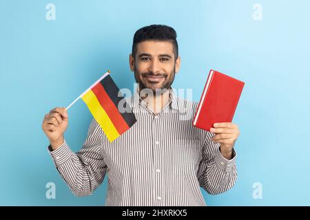 Portrait of positive smiling man showing holding book and german flag, dreaming to study in Germany, education abroad, wearing striped shirt. Indoor studio shot isolated on blue background. Stock Photo