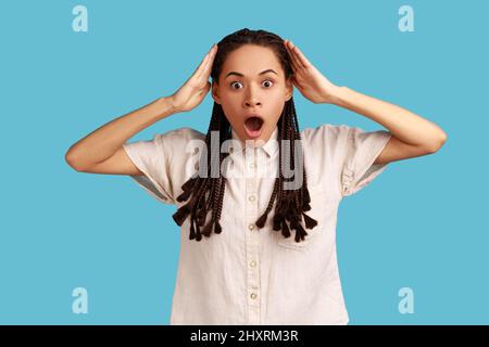 Shocked woman with black dreadlocks reacts on something astonishing, raises hands, hears surprise news, being amazed of something, wearing white shirt. Indoor studio shot isolated on blue background. Stock Photo