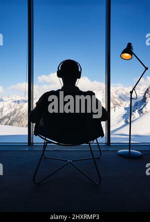 Man in silhouette sitting the chair listen music by headphone near panoramic window with peaceful view of a beautiful snowy mountain landscape. Loft I Stock Photo