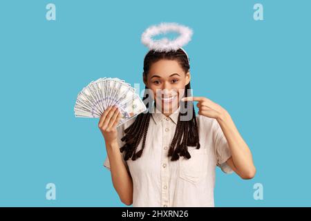 Portrait of smiling angelic woman with dreadlocks and nimb over head holding and pointing at fan of dollars banknotes, wearing white shirt. Indoor studio shot isolated on blue background. Stock Photo