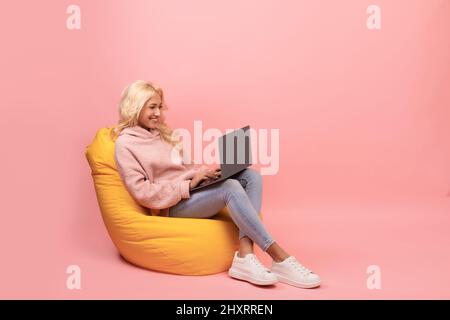 Freelance career. Happy young woman working with laptop computer, sitting in beanbag chair, pink background, copy space Stock Photo