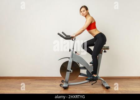 Side view portrait of slim athletic woman with perfect body shape training on bicycle, using sport equipment for workout, wearing sports tights and top. Indoor studio shot on gray wall background. Stock Photo