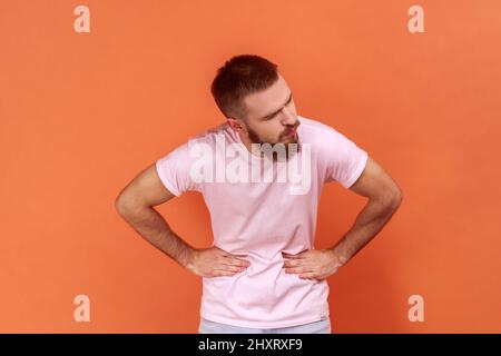 Portrait of sick unhealthy bearded man grimacing and suffering from pain in belly, severe abdominal distress, wearing pink T-shirt. Indoor studio shot isolated on orange background. Stock Photo