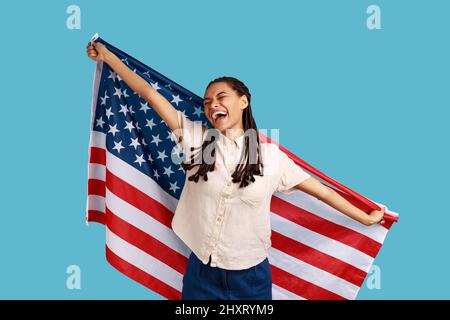 Cheerful attractive woman with black dreadlocks holding USA flag over shoulders and keeps eyes closed and smiling happily, wearing white shirt. Indoor studio shot isolated on blue background. Stock Photo