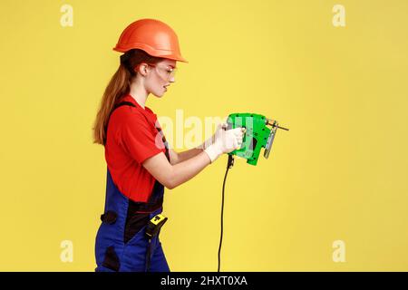 Side view portrait of builder woman working with fretsaw, expressing serious emotions and confidence, wearing overalls and protective helmet. Indoor studio shot isolated on yellow background. Stock Photo