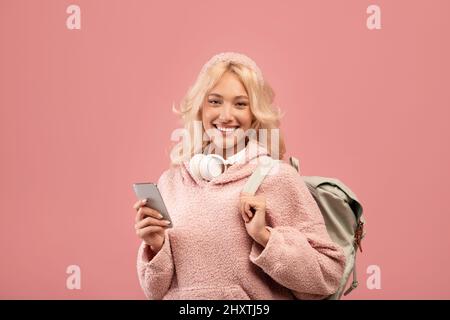 Excited lady student using smartphone and smiling at camera while standing with backpack over pink background Stock Photo