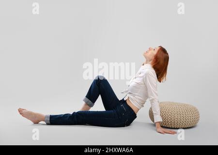 Barefoot middle age woman leaning on padded stool in profile. Studio concept of carefree life. Stock Photo