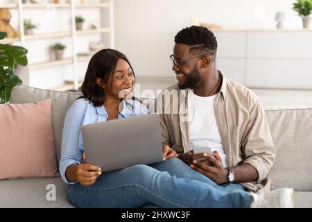 Happy black couple sitting on couch, using cellphone and pc Stock Photo