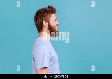 Side view of bearded man looking at camera with toothy smile and happy facial expression, being in good mood, rejoicing great results. Indoor studio shot isolated on blue background. Stock Photo