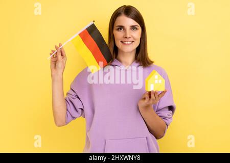 Real estate purchase in Germany, immigration. Portrait of positive woman holding german flag and paper house, wearing purple hoodie. Indoor studio shot isolated on yellow background. Stock Photo