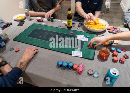 Male friends playing Texas Hold 'em Poker on a table at home with gambling chips, a poker mat, chips and salsa sauce and drinking beers. England Stock Photo