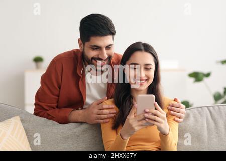 Happy Arabic Spouses Using Phone Together Texting Sitting At Home Stock Photo