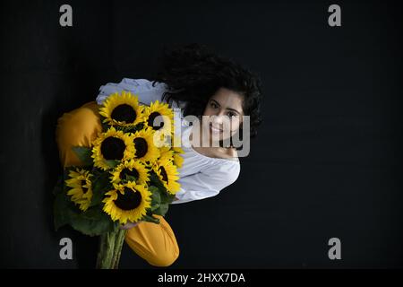 Vertical shot of a Hispanic female posing with sunflowers in front of a black background Stock Photo