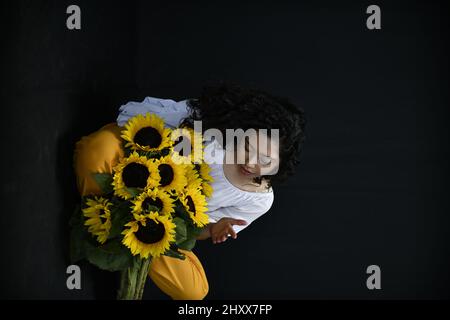Vertical shot of a Hispanic female posing with sunflowers in front of a black background Stock Photo