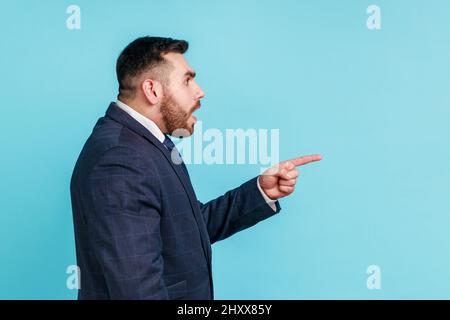 Portrait of attentive handsome bearded young man wearing official style suit having shocked facial expression, pointing finger aside. Indoor studio shot isolated on blue background. Stock Photo