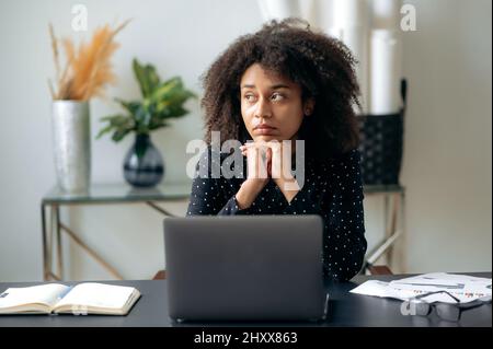 Tired african american curly haired woman, business lady, director, manager, sits in front of a laptop at her desk, rests from computer work, looks to the side, dreams of rest or vacation Stock Photo