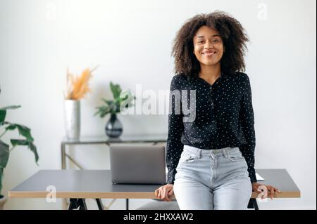 Successful happy pretty young dark-skinned girl, business woman, broker, financial consultant, stands near the desktop in the office, dressed in stylish clothes, looks at the camera, smiles friendly. Stock Photo