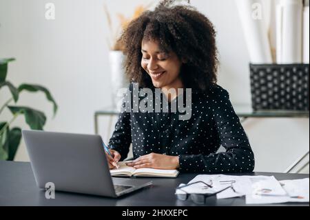 African american company manager or freelancer working in the office. Attractive confident smart curly haired woman, sitting in front of a laptop, making notes in a notebook, listens webinar, smiling Stock Photo