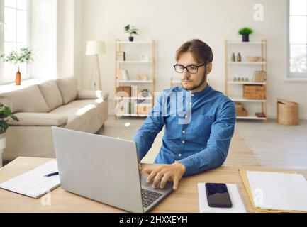 Serious young man sitting at his desk in home office and working on laptop computer Stock Photo