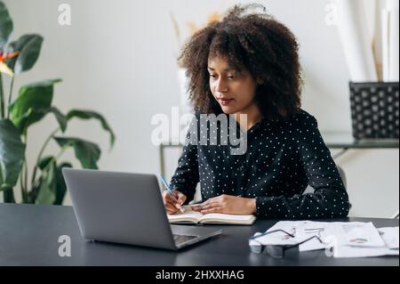 Young adult African American business woman in the office. Attractive confident smart woman top manager or trader, sits in front of a laptop, looks the screen, makes working notes in a notebook Stock Photo