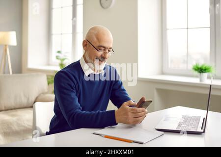 Happy senior man using his mobile phone while sitting at table with laptop computer Stock Photo