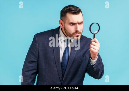 Serious man with beard wearing in dark suit looking away through magnifying glass, spying, finding out something, exploring crime scene, inspecting. Indoor studio shot isolated on blue background. Stock Photo