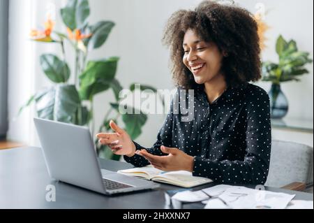 Successful positive young adult woman african woman freelancer, manager, CEO, sitting in office at laptop, talking on video call with client or employees, discussing business strategy, gesturing,smile Stock Photo
