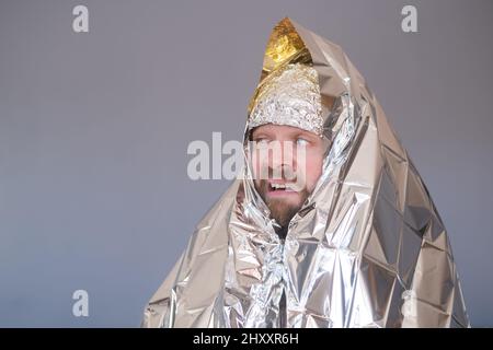 Lost hiker wrapped in an emergency survival blanket, isolated on blue wall. Stock Photo