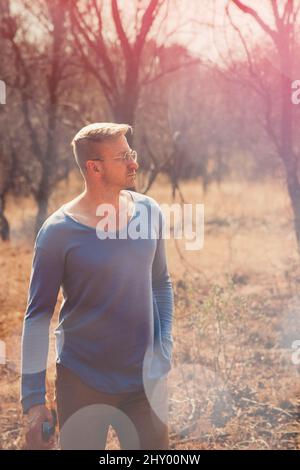 Vertical shot of a young caucasian man with a blue shirt standing in the forest looking to the right Stock Photo