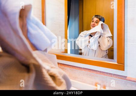 Middle aged female in bathrobe looking at mirror and wiping face during daily morning routine in light bathroom at home Stock Photo