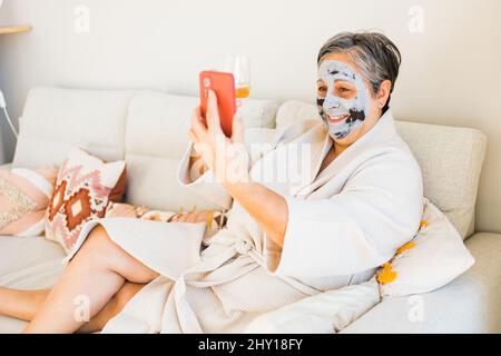 Positive female with clay mask taking selfie while sitting on couch with glass of wine during skincare procedure in living room Stock Photo