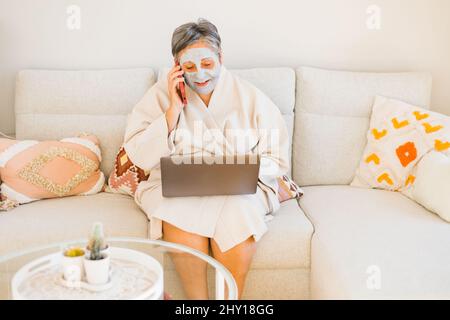 Woman in clay mask on face browsing netbook while sitting on comfortable couch and talking phone in light living room during skincare routine Stock Photo
